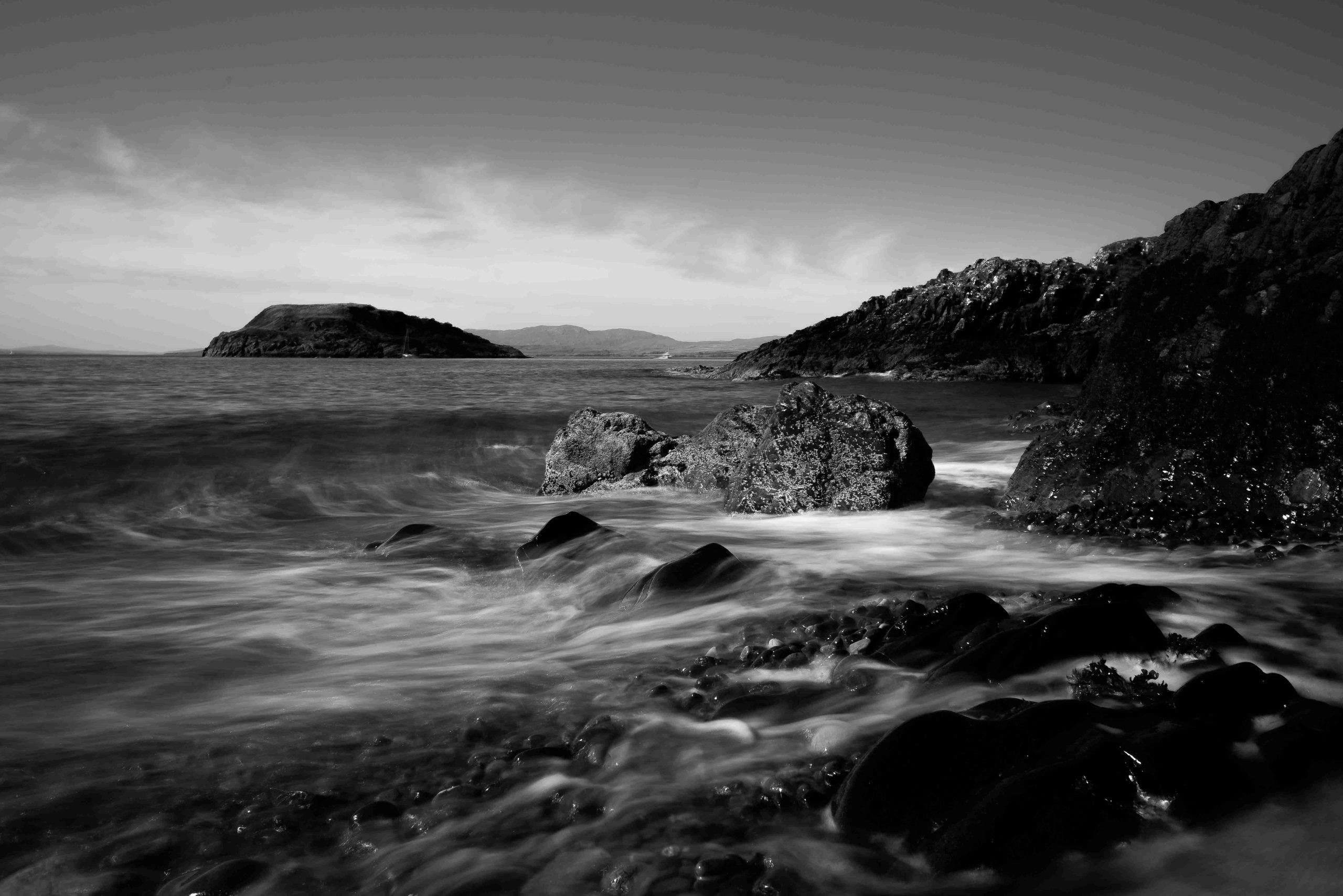 Black and white photograph of waves crashing on a rocky beach, with islands in the background